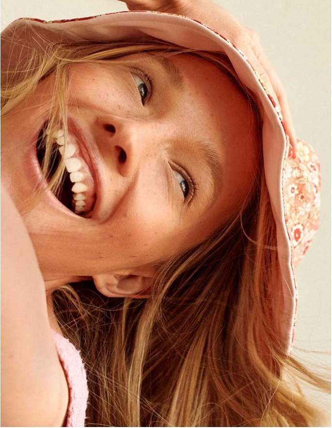 Close-up of a smiling woman with blond hair wearing a patterned bucket hat looking directly at the camera in a bright indoor setting.