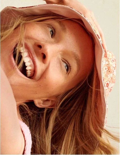 Close-up of a smiling woman with blond hair wearing a patterned bucket hat looking directly at the camera in a bright indoor setting.