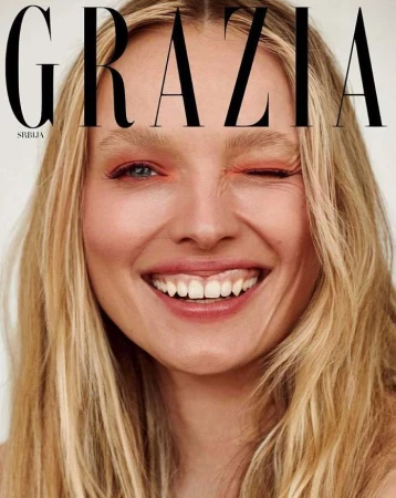 Close-up portrait of a blonde woman smiling and winking with orange eyeshadow against a light background.
