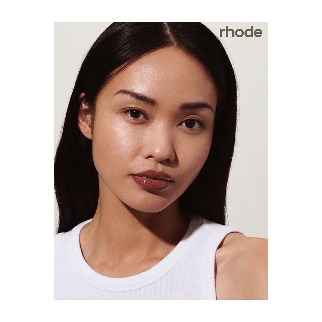 Close-up studio shot of an adult woman with dark hair, wearing a white top, and posing in front of a neutral background with Rhode branding.