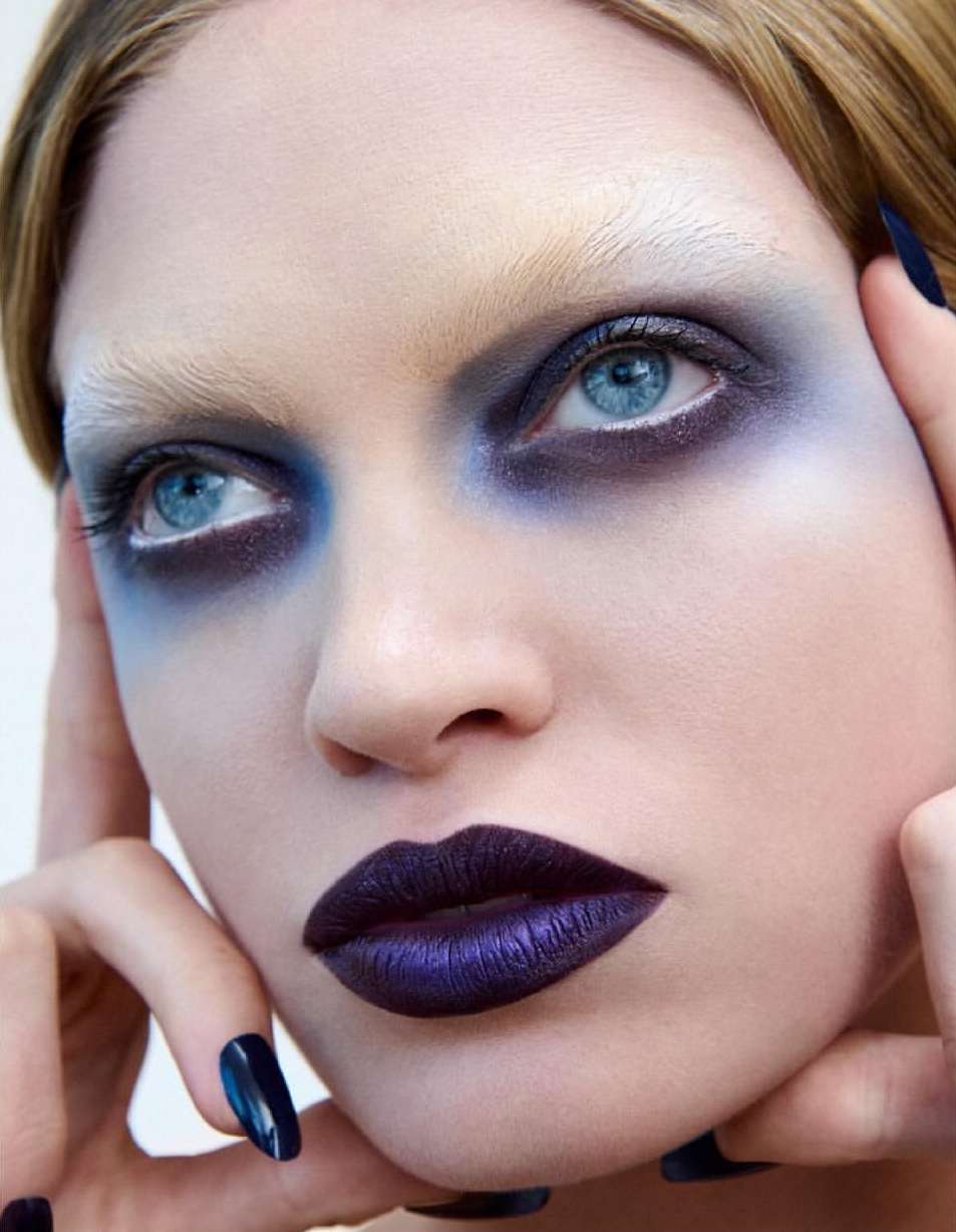 Close-up of a woman with striking blue eyeshadow, white eyebrows, and dark purple lipstick, creating a bold beauty statement.