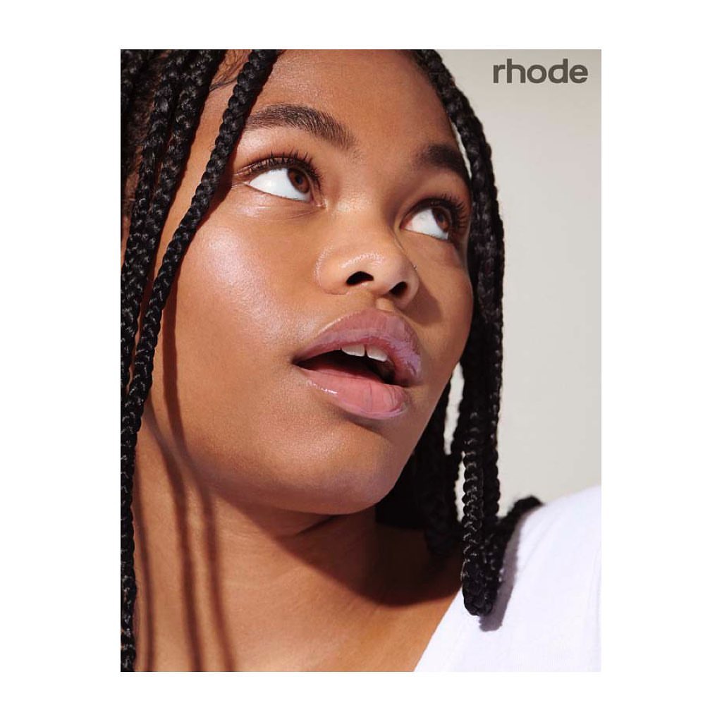 Close-up portrait of a young Black woman with braided hair and glossy lips looking upward against a plain white background.