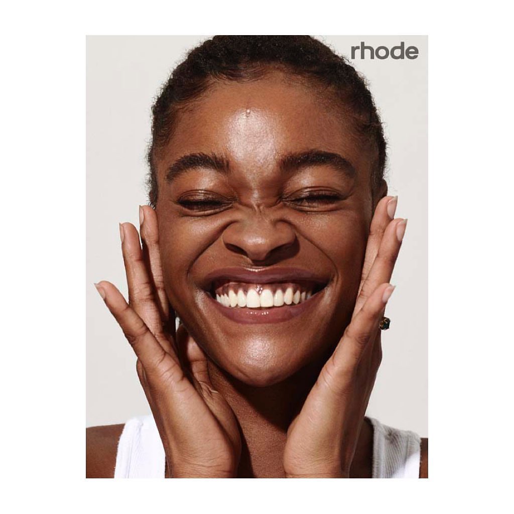 Close-up of a smiling dark-skinned woman with her eyes closed and hands on her cheeks. The woman is wearing a white tank top.