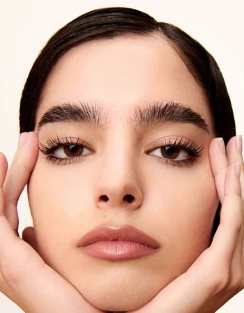 Close-up beauty portrait of a young woman framing her face with both hands, featuring defined dark eyebrows and neutral lipstick