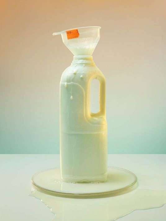 Still life studio shot of a white plastic milk jug with a funnel, overflowing with liquid, sitting on a reflective surface.