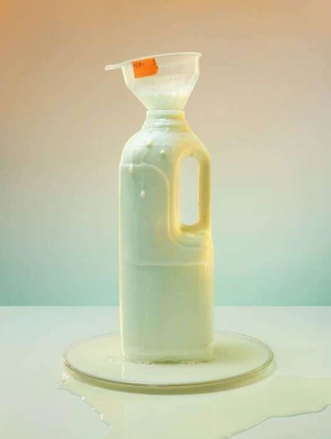 Still life studio shot of a white plastic milk jug with a funnel, overflowing with liquid, sitting on a reflective surface.