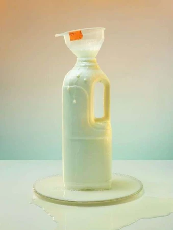 Still life studio shot of a white plastic milk jug with a funnel, overflowing with liquid, sitting on a reflective surface.