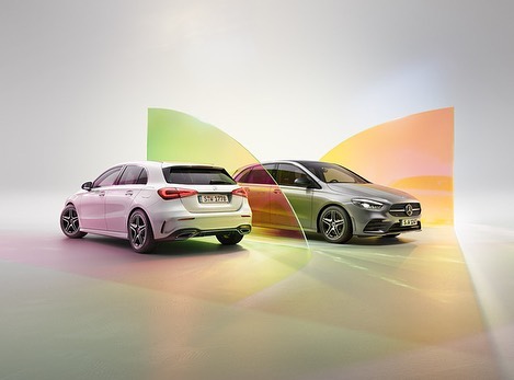 Two Mercedes-Benz cars, a white hatchback and a silver sedan, are displayed in a studio setting with colorful geometric lights.