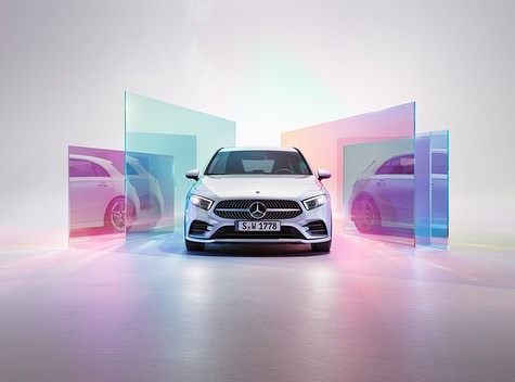 Silver Mercedes-Benz A-Class hatchback in a studio, set against a backdrop of translucent panels with iridescent color effects.