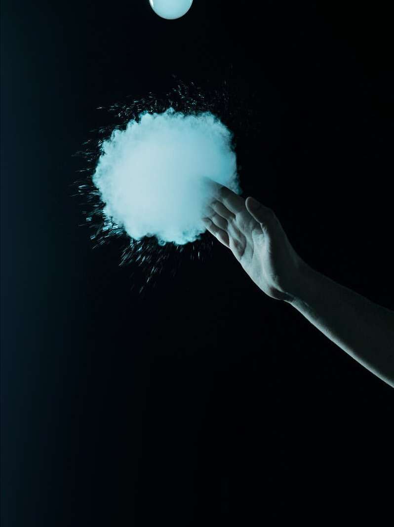 Hand reaches toward a cloud of vaporous material in a dark space, illuminated by a glowing orb above. Moody lighting and dark background.