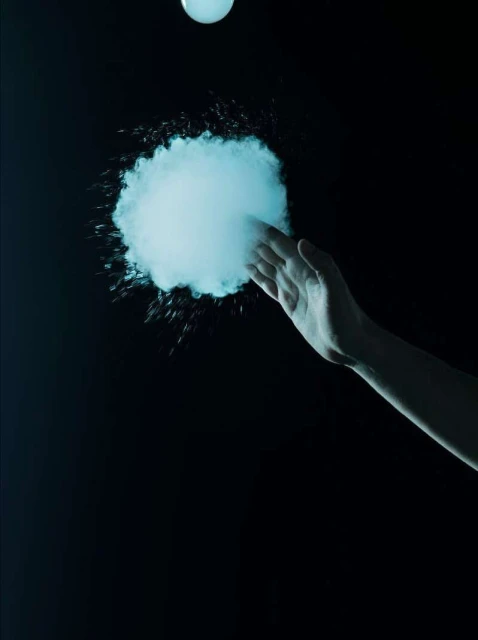 Hand reaches toward a cloud of vaporous material in a dark space, illuminated by a glowing orb above. Moody lighting and dark background.