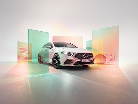 White Mercedes-Benz A-Class hatchback car parked indoors against a white background with glass panels reflecting colorful light.