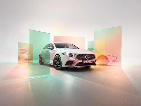 White Mercedes-Benz A-Class hatchback car parked indoors against a white background with glass panels reflecting colorful light.