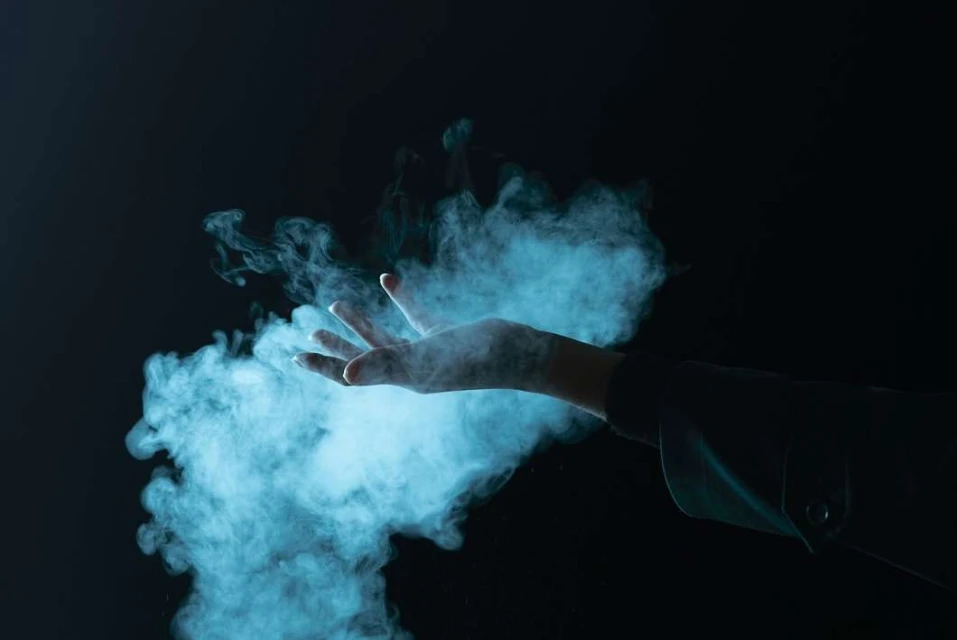 Hand reaching into a swirling cloud of light blue smoke against a dark background, creating a mysterious and ethereal atmosphere.