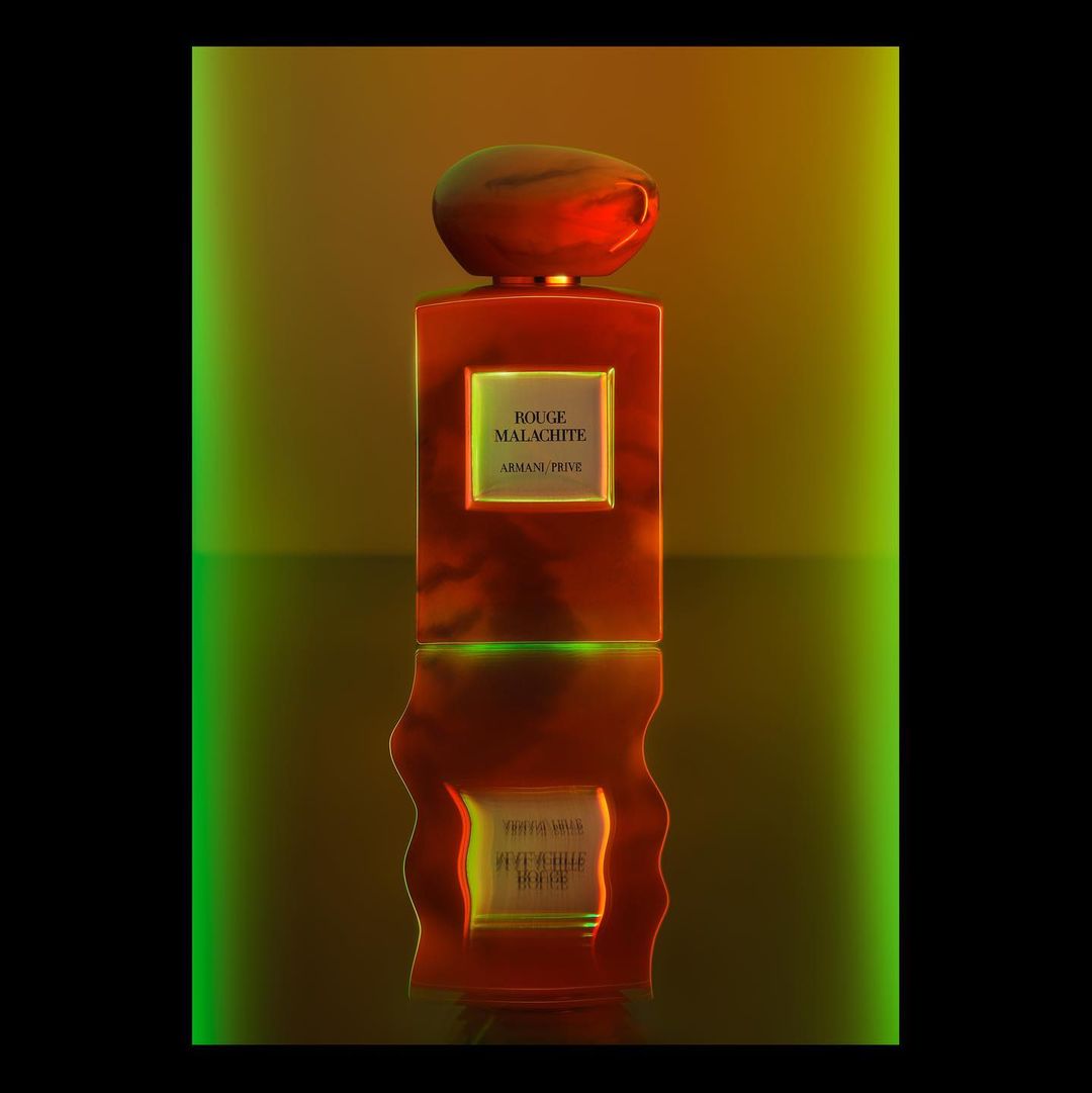 A red glass perfume bottle with a rounded cap and the text 'Rouge Malachite Armani Prive' is centered on a reflective surface against green and gold.