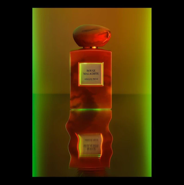 A red glass perfume bottle with a rounded cap and the text 'Rouge Malachite Armani Prive' is centered on a reflective surface against green and gold.
