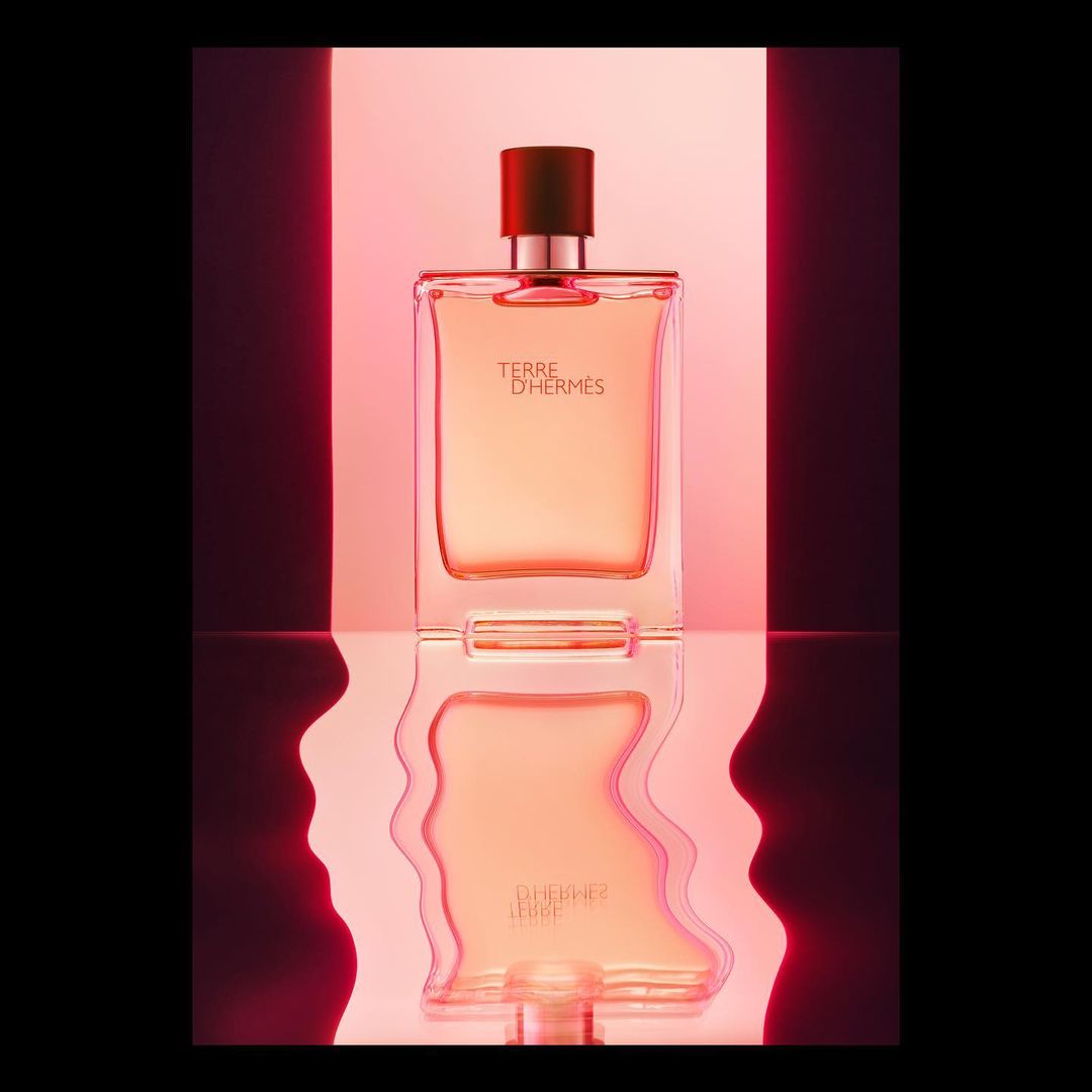 Perfume bottle on a reflective surface with pink and red lighting. Terre D'Hermes bottle reflecting on a glossy plane, studio lighting.
