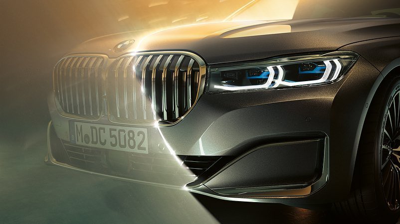 Close-up of a grey BMW car featuring its distinctive grill, modern headlights, and the hood. The vehicle is illuminated by golden lighting.