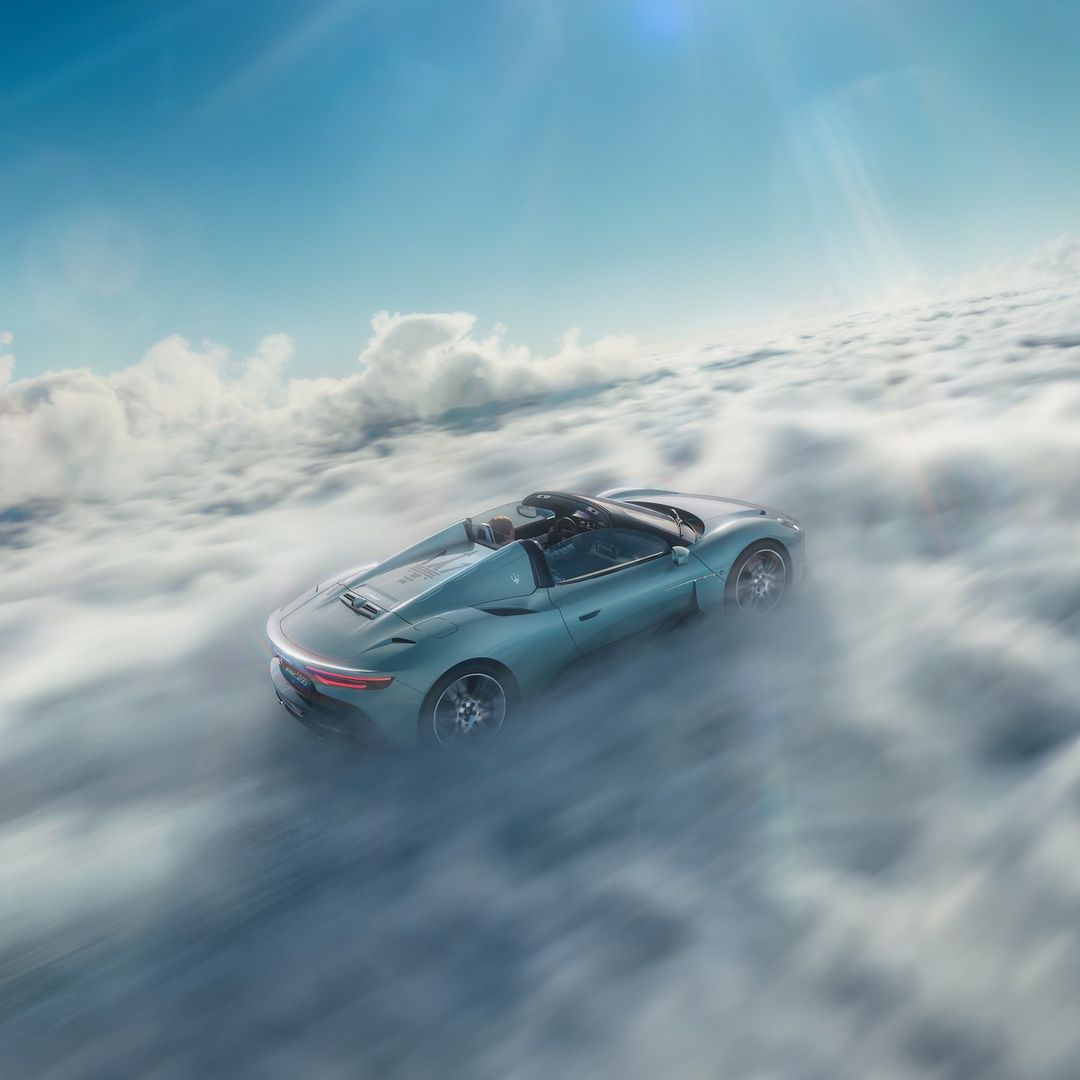 A light blue convertible car drives through a thick cloudscape under a blue sky, creating a dynamic and surreal automotive image.