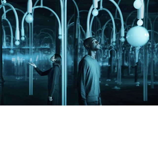 Young adults inside an abstract light display room with spherical bulbs and light emitting from objects. The scene is dark and moody.