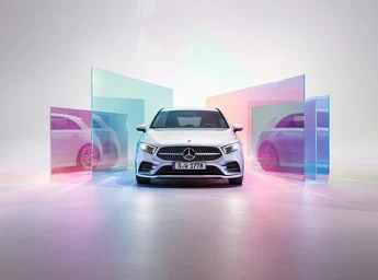 Front view of a white car parked in a minimalist studio surrounded by translucent colored glass panels reflecting pink and green hues.