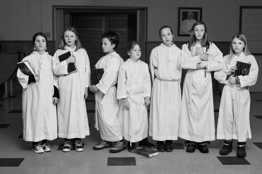 Black-and-white photo of seven children in white robes holding books, standing in a row in a church interior with tiled floors.