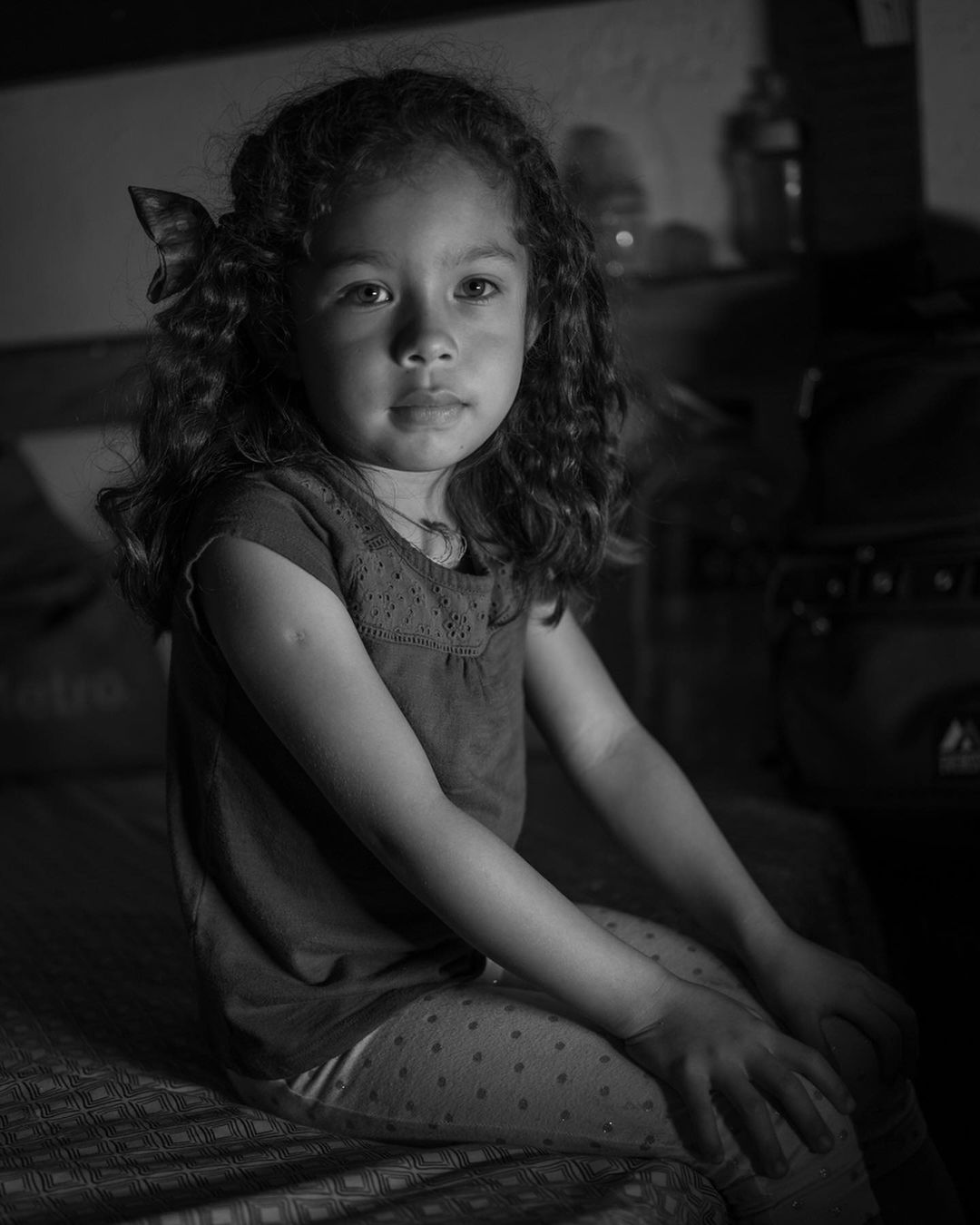 Monochrome portrait of a child with curly hair sitting indoors looking at the camera with a soft, diffused light illuminating her face.