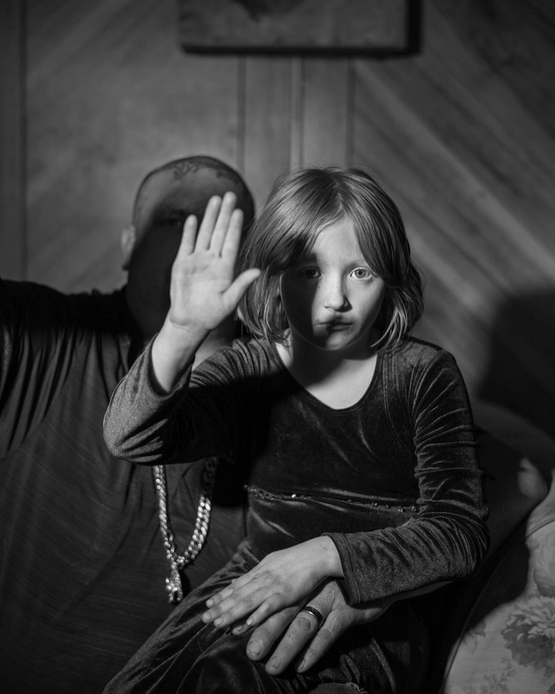 Black and white portrait of a girl with hand up, sitting with an adult with visible jewelry indoors with wood paneling backdrop.