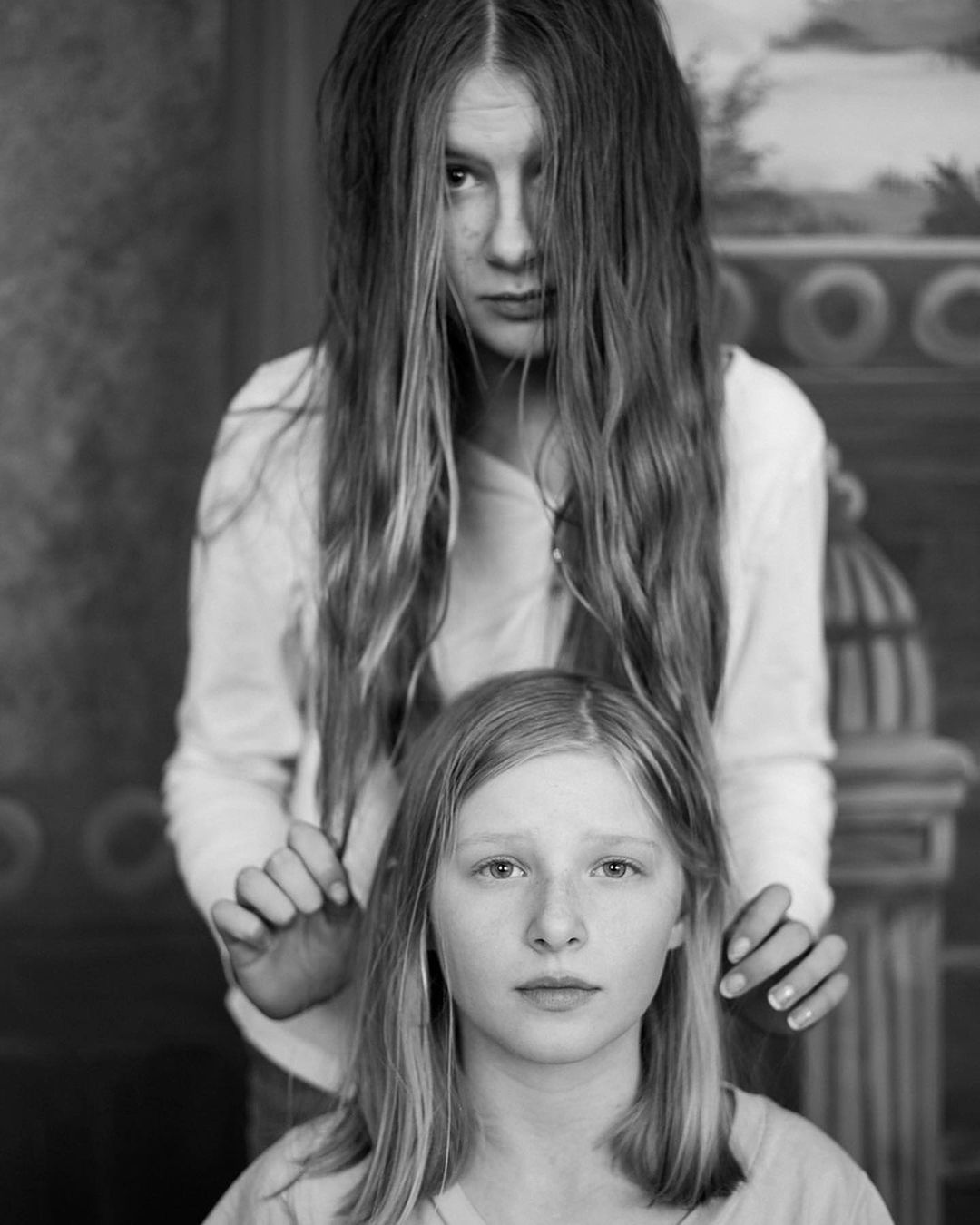 Black-and-white portrait of two young women in casual clothing, one standing behind the other against a backdrop with artistic painting.