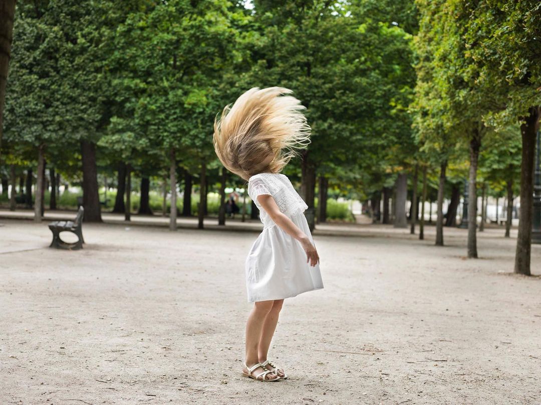 Young girl in a white dress flicks her long blonde hair in a city park with benches and trees, creating a sense of youthful energy and freedom.