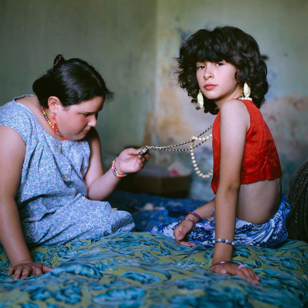 Two young girls indoors are seated on a patterned bed; one adjusts the other's necklace, both wearing large earrings and bracelets.
