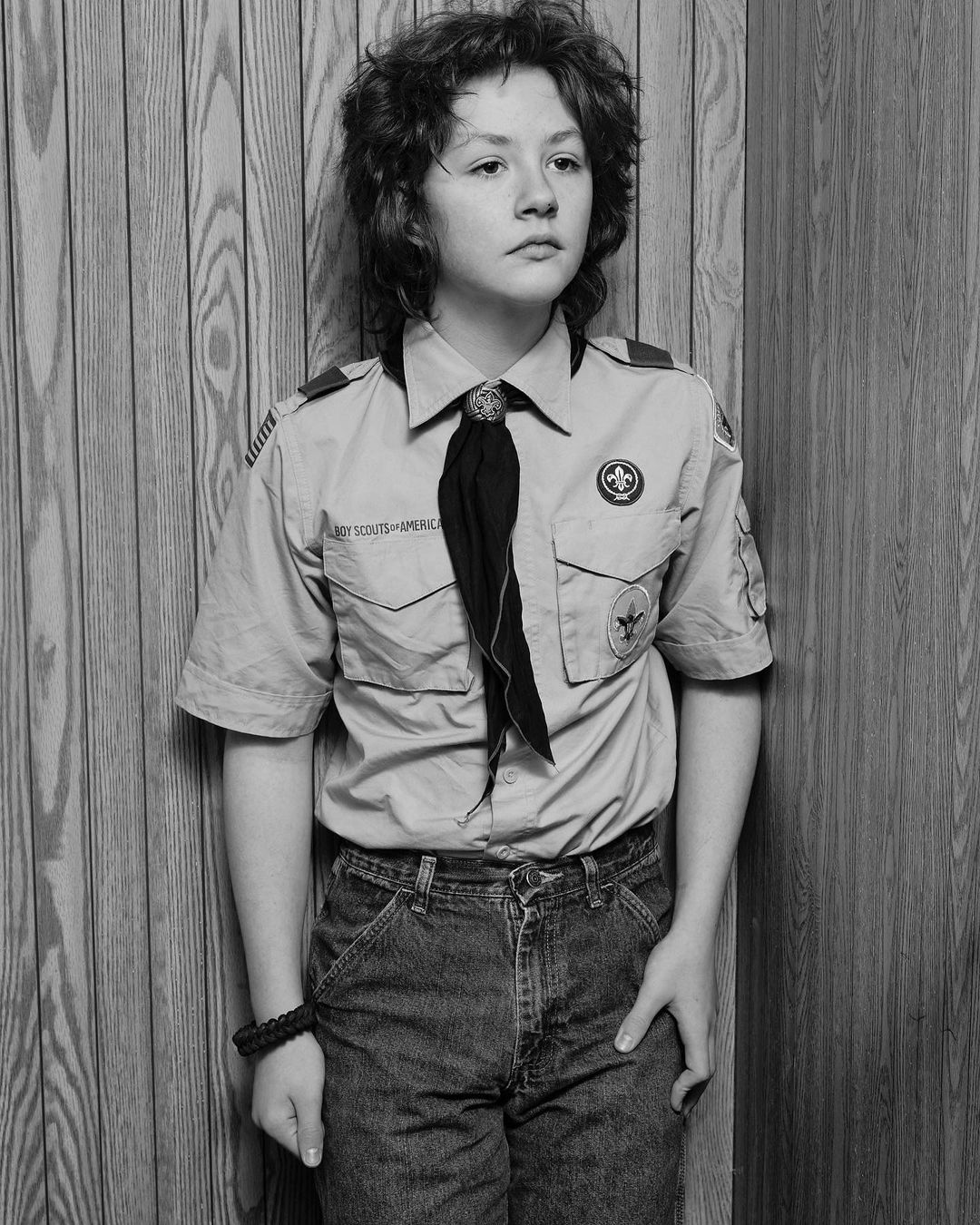 Black and white portrait of a young person in a Boy Scouts of America uniform standing against a wood-paneled wall with neutral expression.