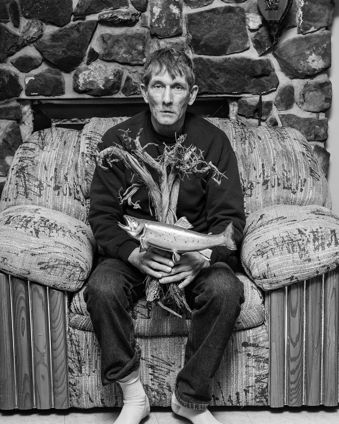 Monochrome portrait of a person sitting on a floral couch indoors, holding a wooden fish and a bunch of sticks in front of a stone fireplace.