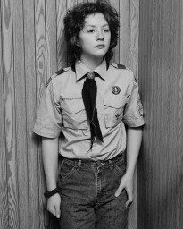Black and white portrait of a young person wearing a scout uniform and denim jeans against a wood-paneled wall.