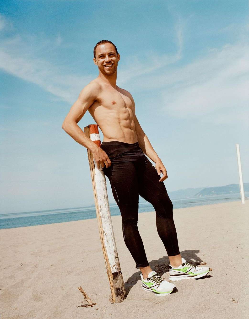 Athletic man resting against a wooden post at the beach wearing black leggings and sneakers, with a blue sky and ocean backdrop.