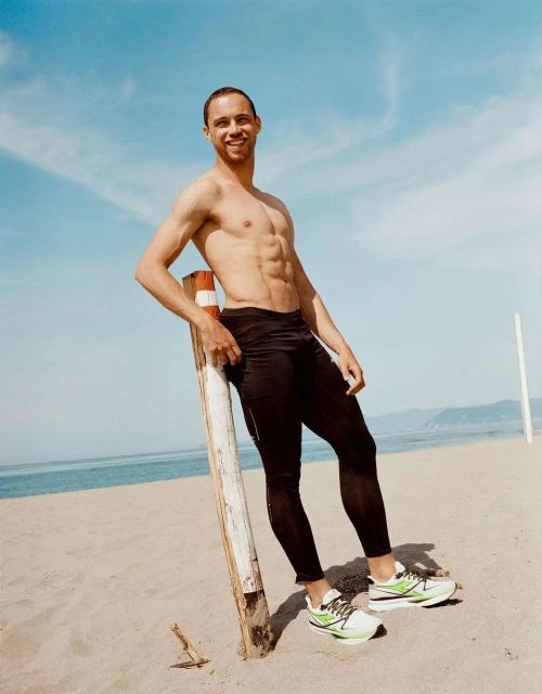Athletic man resting against a wooden post at the beach wearing black leggings and sneakers, with a blue sky and ocean backdrop.