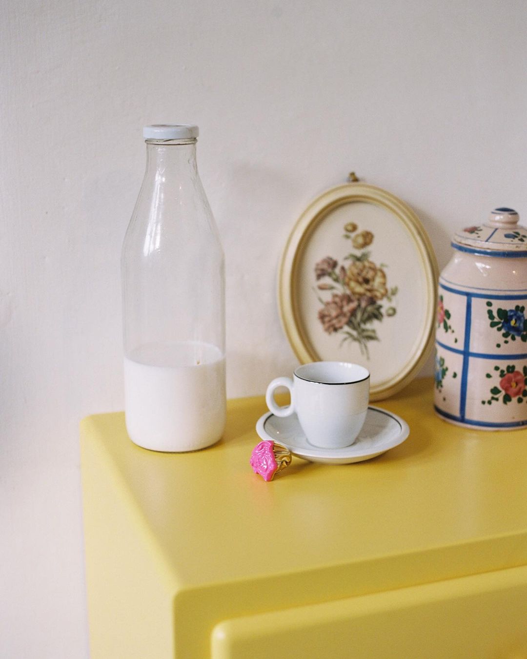Still life of a milk bottle, tea cup, candy ring, painting, and ceramic jar on a yellow dresser against a cream wall.