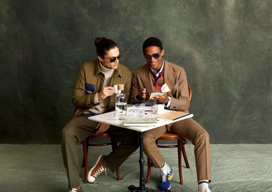 Two men sitting at a table, one holding a book and the other a coffee cup, both wearing sunglasses, in an indoor setting with drinks.