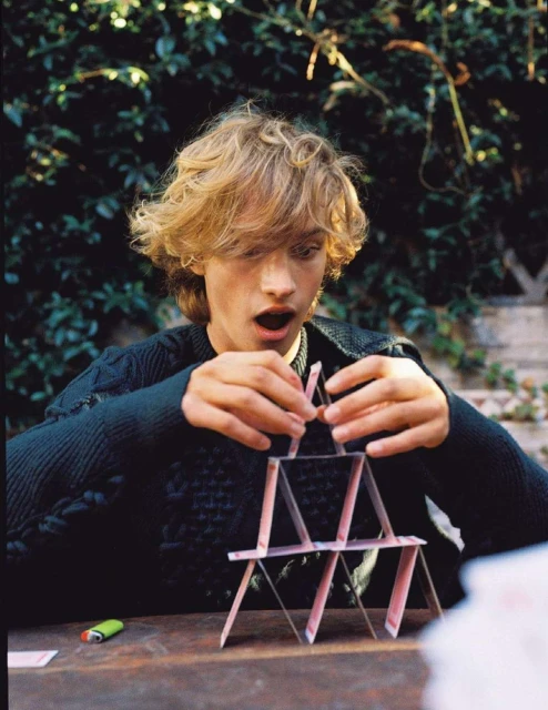 Young adult with blond hair concentrating on building a playing card tower indoors on a wooden table with a leafy background.