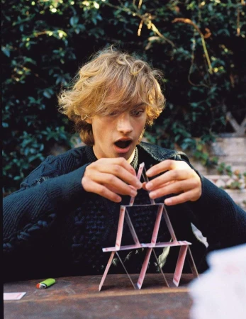 Young adult with blond hair concentrating on building a playing card tower indoors on a wooden table with a leafy background.
