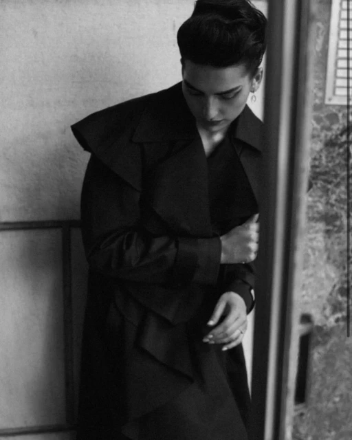Monochrome portrait of a woman with sleek hair wearing a trench coat, set indoors near a window. Fashion editorial photography.