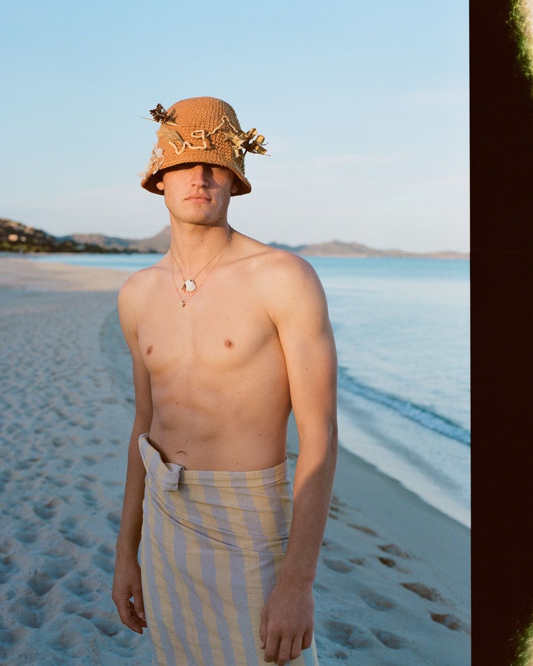 Portrait of a shirtless man on a sandy beach wearing a patterned bucket hat and a yellow and white striped sarong-style wrap.