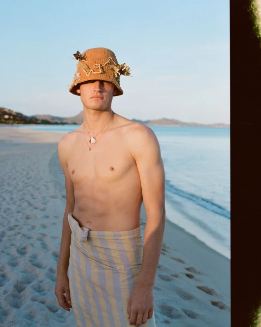 Portrait of a shirtless man on a sandy beach wearing a patterned bucket hat and a yellow and white striped sarong-style wrap.