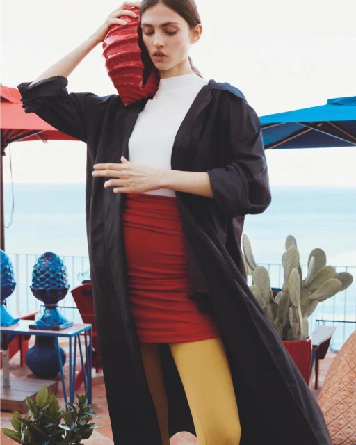 A woman poses outdoors in a black coat, red skirt, and yellow tights, holding a red vase against a backdrop of the sea and sky.