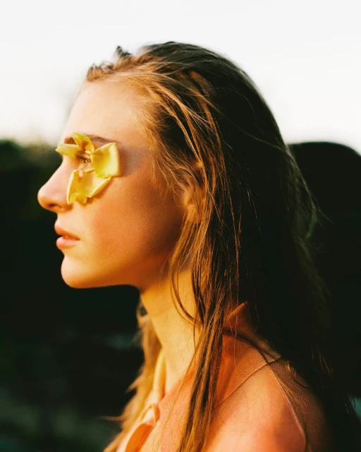 Profile of woman's face with blonde hair and yellow flower petals over her eye, soft lighting, and a dark blurred background.