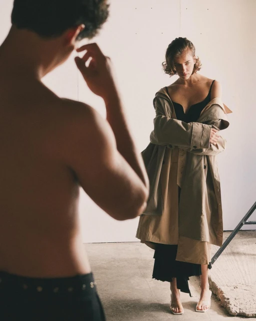 Fashion shot of a woman in a coat and underwear, standing near a topless man in pants against a white background in a studio.