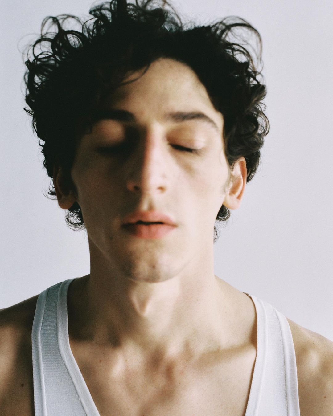 Close-up portrait of a young adult male with dark curly hair and closed eyes, wearing a white ribbed tank top against a plain background.