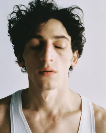 Close-up portrait of a young man with dark curly hair and closed eyes wearing a white ribbed tank top.