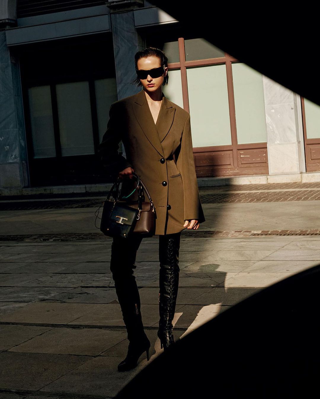 Model wearing dark sunglasses, brown blazer, and tall black boots standing on a stone street holding a handbag