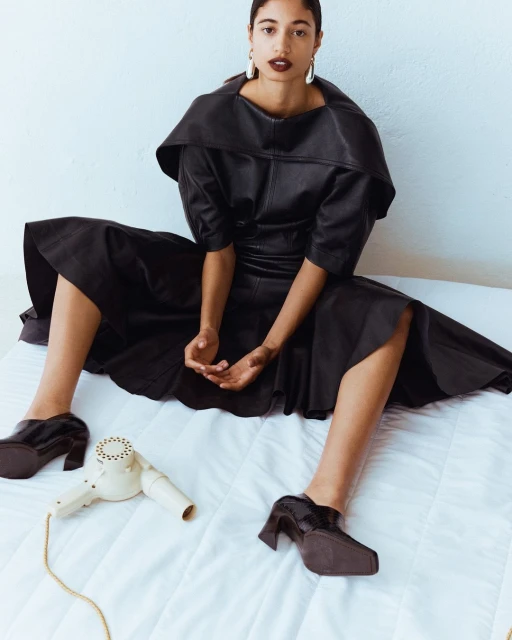 Woman in a black dress and shoes sitting on a bed next to a hair dryer with white walls. Fashion, beauty, and lifestyle portrait.